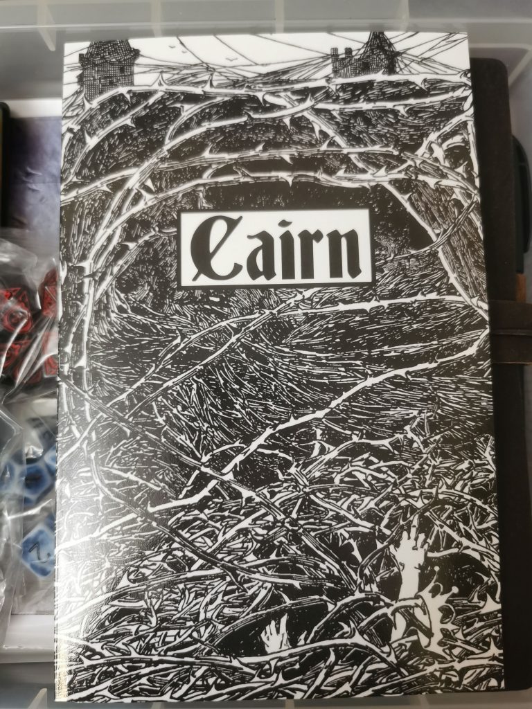 A photograph of the black and white cover of the RPG CAIRN, lying on top of a box of dice and other gaming stuff.